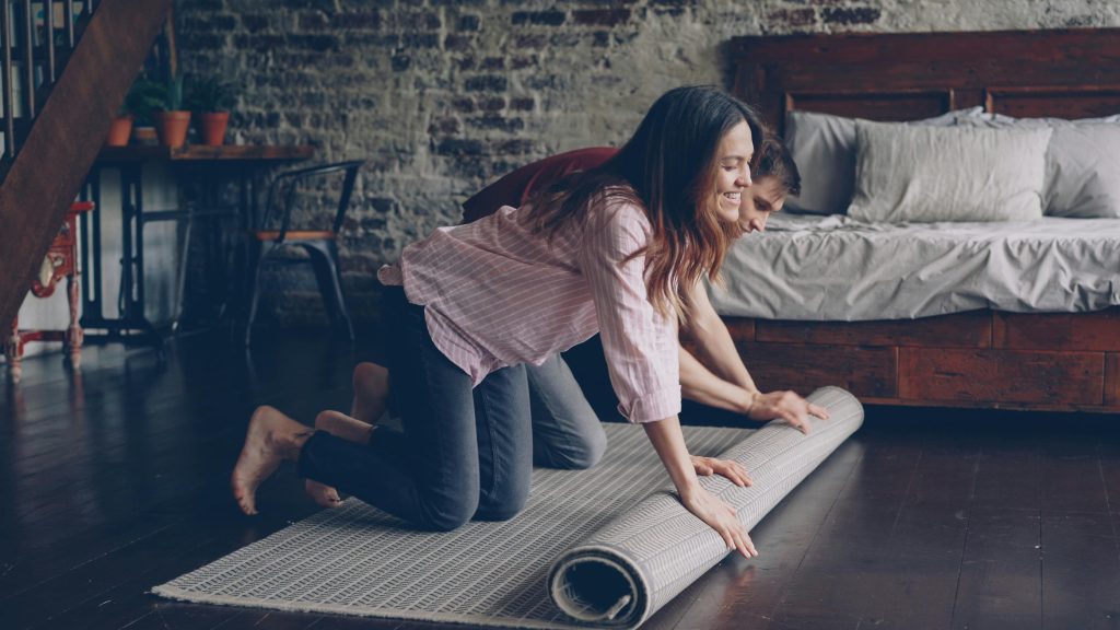 A happy couple rolling a rug on a wooden floor in a comfortable, rustic bedroom.