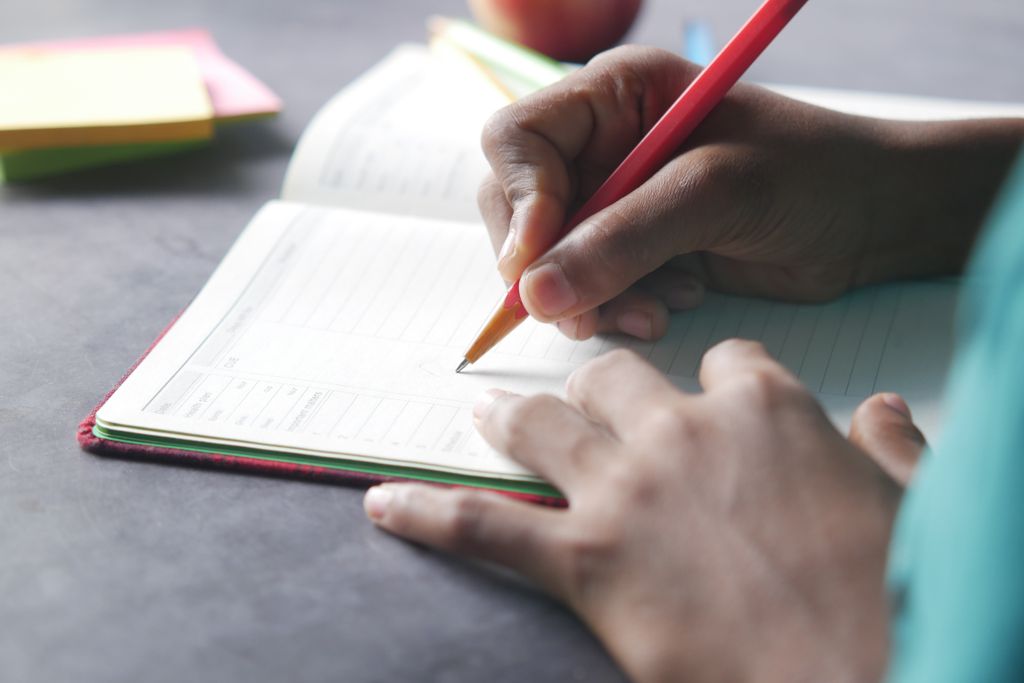 Close-up of a hand writing in a notebook with a pencil indoors.
