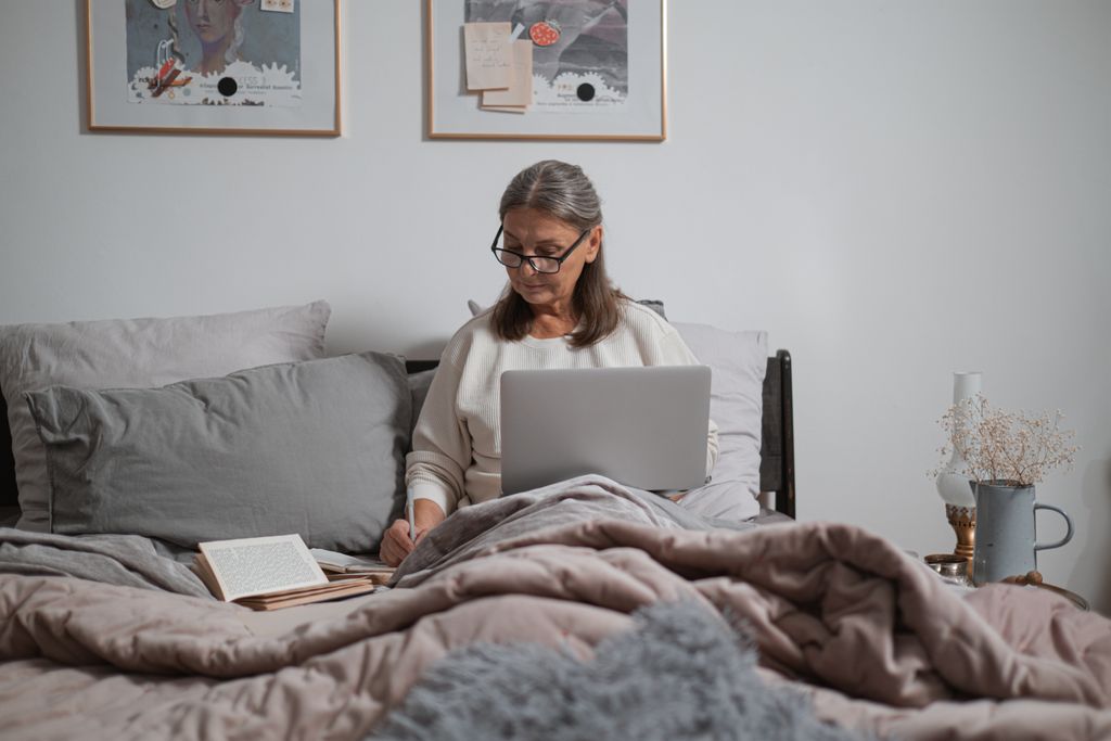 Elderly woman sitting on bed using laptop, writing notes. Comfortable home setting.