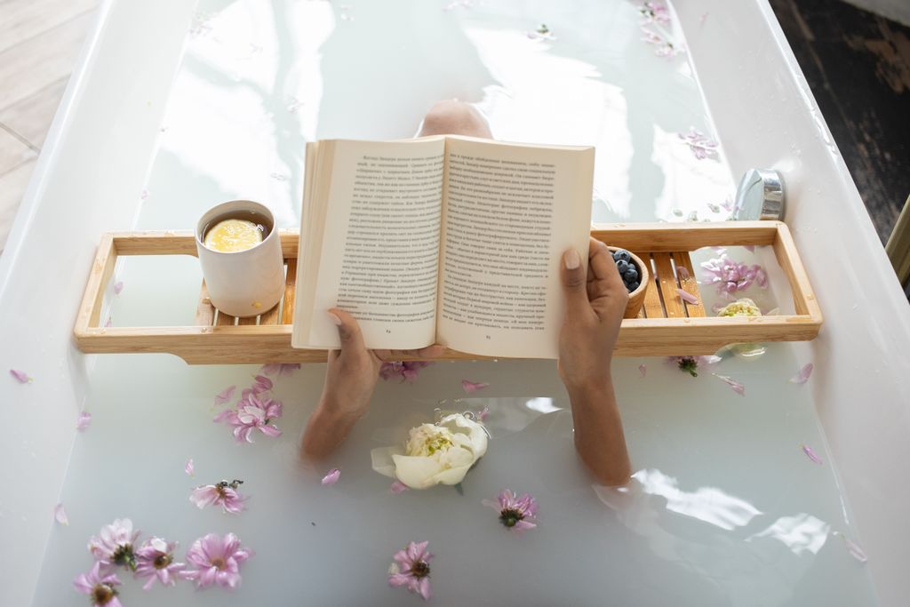 From above of anonymous female reading book while taking bath in water with white and pink flowers