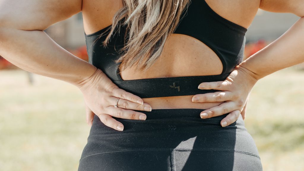 A woman in activewear holds her lower back, indicating muscle strain, outdoors.